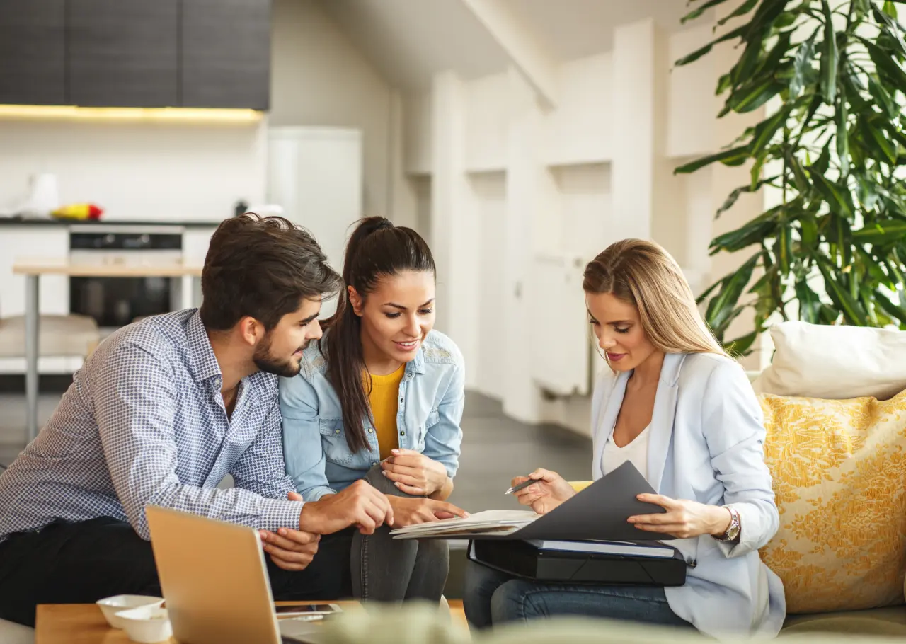 Three Expert Realtors in an office setting discuss documents, with a laptop open on the table. A large plant adds a touch of greenery in the background, creating a lively and professional atmosphere.