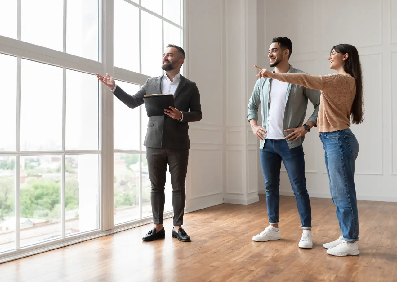 A realtor shows a spacious, bright apartment to a smiling man and woman. They are looking out the large windows at the view.