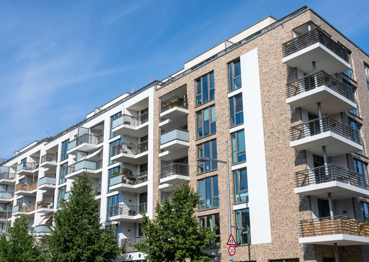 Modern apartment building with brick and white facade, large windows, and balconies. Trees and a traffic sign are visible in the foreground under a clear blue sky.