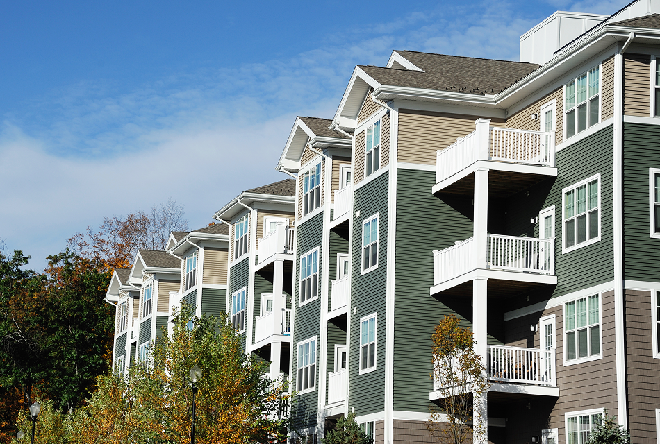 Modern apartment complex with a green and beige exterior designed by real estate experts, featuring multiple balconies and white trim against a clear blue sky, with trees beautifully framing the view.