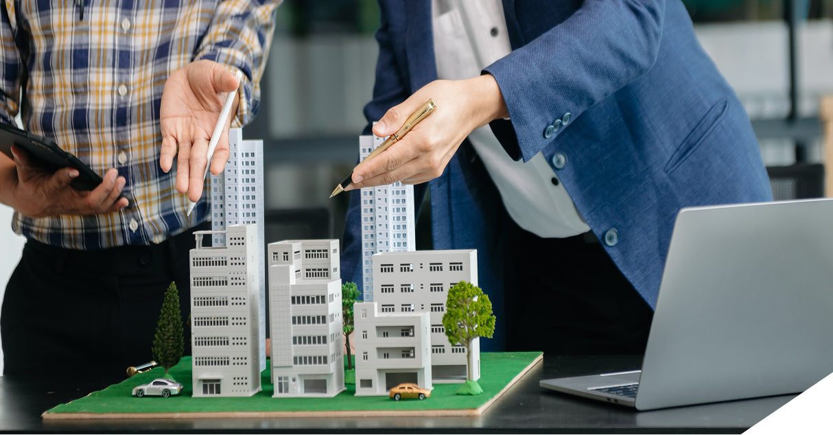 Two real estate experts examine a city model with buildings and trees on a table; one holds a tablet, the other gestures with a pen, as a laptop sits open nearby.