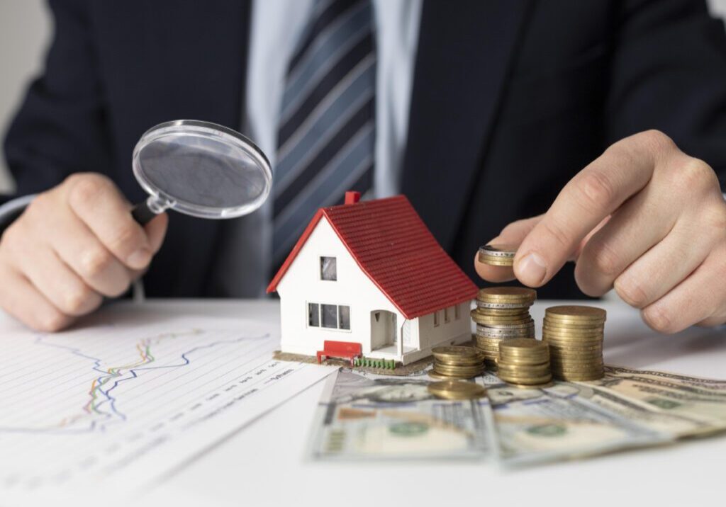 A person in a suit examines a miniature house with a magnifying glass while stacking coins and counting dollar bills on a table with financial charts.